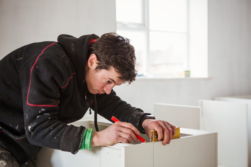 A builder, holding a pencil and ruler, measuring a kitchen unit.